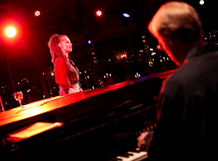 Sandra Kreisler steht im roten Licht am Flügel, im Vordergrund der Pianist